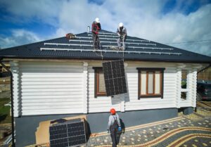 Men mounters installing solar panel system on roof of house. Workers in helmets lifting up photovoltaic solar module with help of ropes outdoors. Concept of alternative and renewable energy.