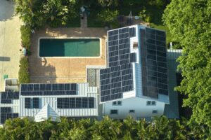 Aerial view of a house with solar panels on the roof, installed by St John the Baptist Solar Installers, next to a rectangular swimming pool surrounded by trees and greenery.