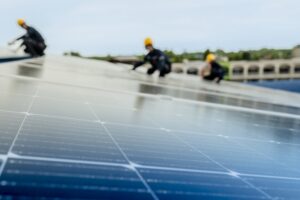 Tangipahoa Parish Solar installers in yellow helmets install or inspect solar panels on a rooftop, with a blurred background of buildings and sky.