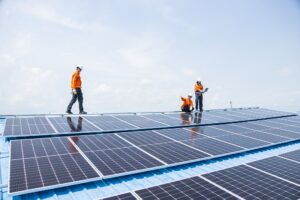 Three workers in orange safety gear and helmets inspect and work on rooftop solar panels under a clear sky, showcasing the expertise of Tangipahoa Parish Solar installers.