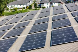 Rows of solar panels are installed on a large, metal rooftop by St John the Baptist Solar Installers, with residential houses visible in the background.