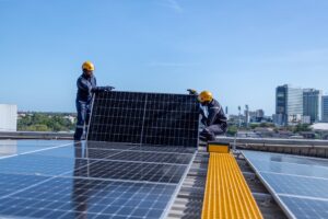 Two workers in safety gear, representing Solar Installers in Plaquemines Parish, install a large solar panel on a rooftop with city buildings visible in the background.