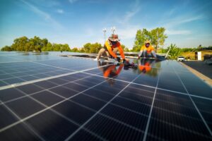 Two workers, trusted solar installers in Plaquemines Parish, install solar panels on a rooftop under a clear blue sky, with trees and equipment visible in the background.