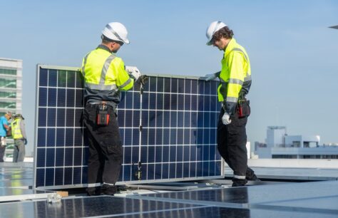 Two technicians in high-visibility jackets and helmets carefully install a solar panel on a rooftop with urban buildings in the background.