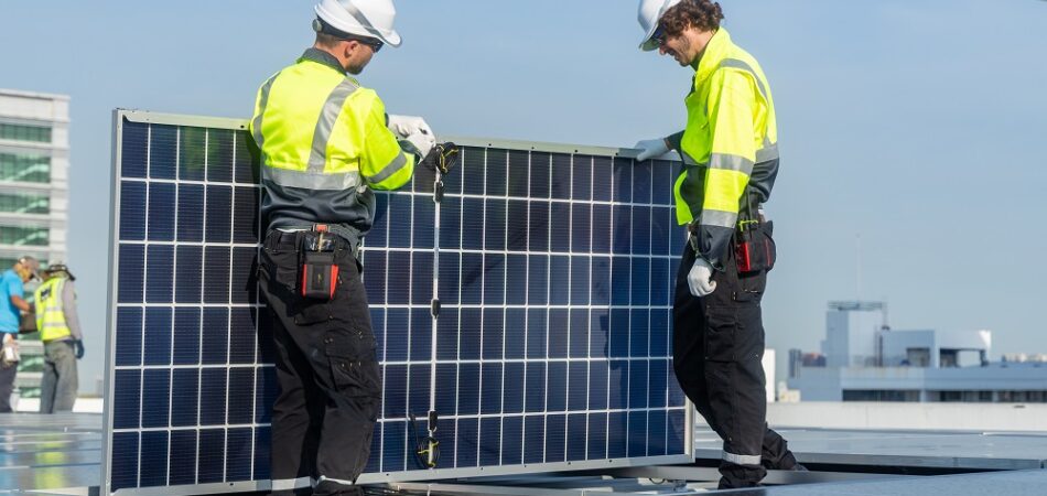 Two technicians in high-visibility jackets and helmets carefully install a solar panel on a rooftop with urban buildings in the background.