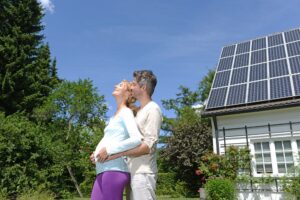 A couple stands outside a house with solar panels on the roof, embracing and looking up on a sunny day—excited for savings thanks to the Federal Solar Tax Credit 2026.
